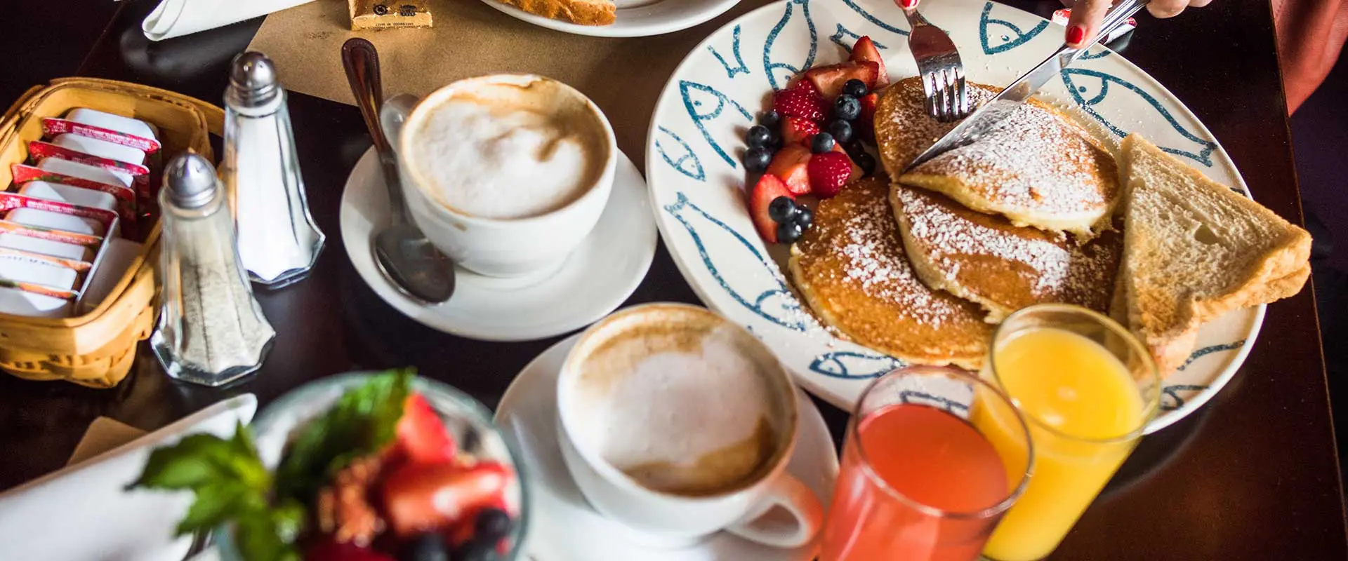 A brunch table set with pancakes dusted in powdered sugar, fresh berries, toast, cappuccinos and juice